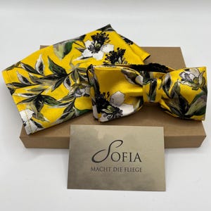May include: A yellow floral bow tie and matching pocket square set. The set features a vibrant yellow background with white and black floral patterns. A business card with the name "Sofia" is also included.