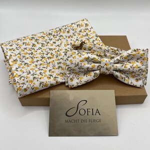 Op de afbeelding: Een witte vlinderdas, pochet en hoofdband set met een geel bloemenpatroon. De set wordt gepresenteerd op een bruine doos, met een visitekaartje met de tekst "Sofia Macht Die Fliege".