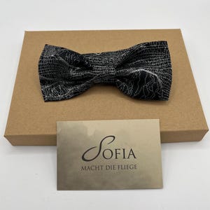 May include: A black and white patterned bow tie rests on a brown cardboard box. A business card with the name "Sofia" and the phrase "Macht die Fliege" is in front of the bow tie. The bow tie has a textured design.
