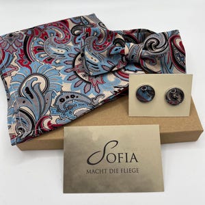 May include: A set of accessories including a paisley patterned bow tie and pocket square in red, blue, and gray. The set includes a pair of round cufflinks and a card with the name "Sofia" on it.
