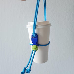 May include: A white paper coffee cup held by a blue cord cup holder. The holder has a blue buckle and a green button. The cord is knotted at the bottom and held by a hand.