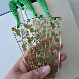 May include: Four rectangular resin bookmarks with gold metal frames, each containing pressed green leaves and gold foil flakes. Each bookmark features a bright green fabric tassel attached to the top through a small circular metal loop.