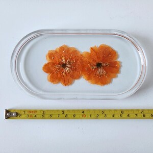 May include: An oval-shaped resin tray with two orange flowers embedded inside. The tray is clear with a light blue background. A yellow tape measure is placed below the tray, showing measurements in inches.