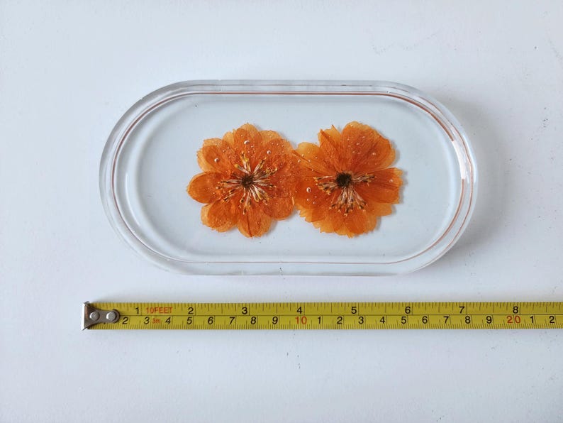 May include: An oval-shaped resin tray with two orange flowers embedded inside. The tray is clear with a light blue background. A yellow tape measure is placed below the tray, showing measurements in inches.