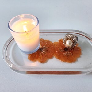 May include: A clear resin tray with a lit candle in a glass holder, two dried orange flowers, and a silver ring with a pearl. The tray is oval-shaped with a copper-colored border. The candle is white and has a soft glow.