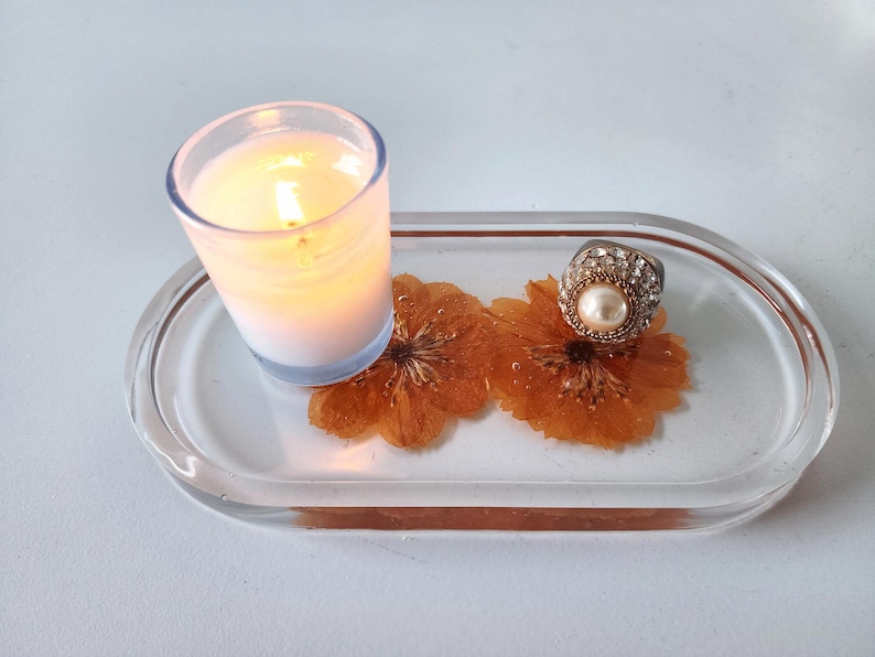 May include: A clear resin tray with a lit candle in a glass holder, two dried orange flowers, and a silver ring with a pearl. The tray is oval-shaped with a copper-colored border. The candle is white and has a soft glow.