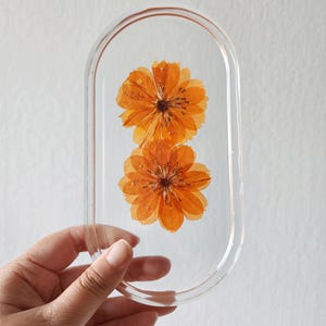 May include: Clear resin oval-shaped tray with two orange flowers encased inside. The flowers have multiple layers of petals and are a vibrant orange color. The tray is held by a hand against a white background.