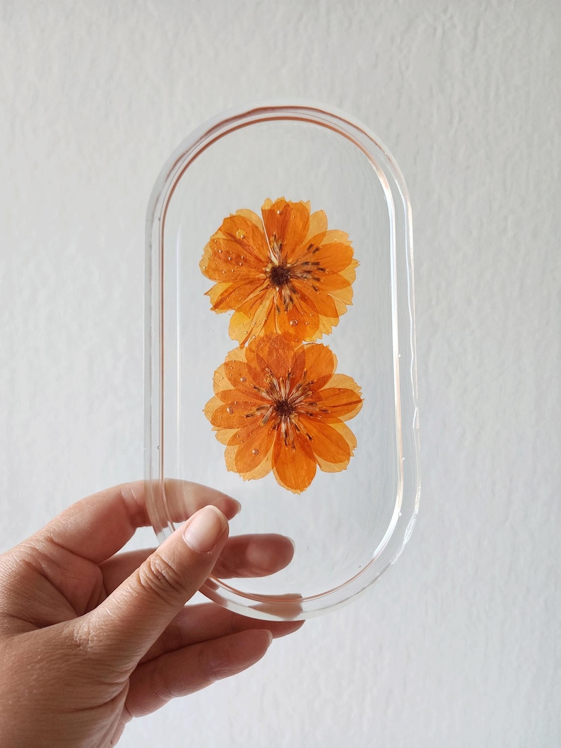 May include: Clear resin oval-shaped tray with two orange flowers encased inside. The flowers have multiple layers of petals and are a vibrant orange color. The tray is held by a hand against a white background.