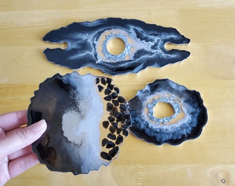 May include: Three resin coasters in black, white, and gold with a glittery finish. Two coasters have a circular shape with a hole in the center, and one coaster is shaped like a wine bottle holder with two holes for the bottle neck.