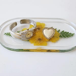 May include: A clear, oval-shaped resin tray with a silver and gold watch-style bracelet, a heart-shaped ring, and yellow flowers and greenery embedded inside. The tray is on a white surface.