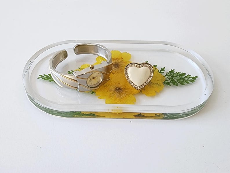 May include: A clear, oval-shaped resin tray with a silver and gold watch-style bracelet, a heart-shaped ring, and yellow flowers and greenery embedded inside. The tray is on a white surface.