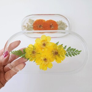 May include: Two clear, oval-shaped resin trays. One tray features orange flowers, while the other holds yellow flowers and green fern-like leaves. The trays are displayed on a white background.
