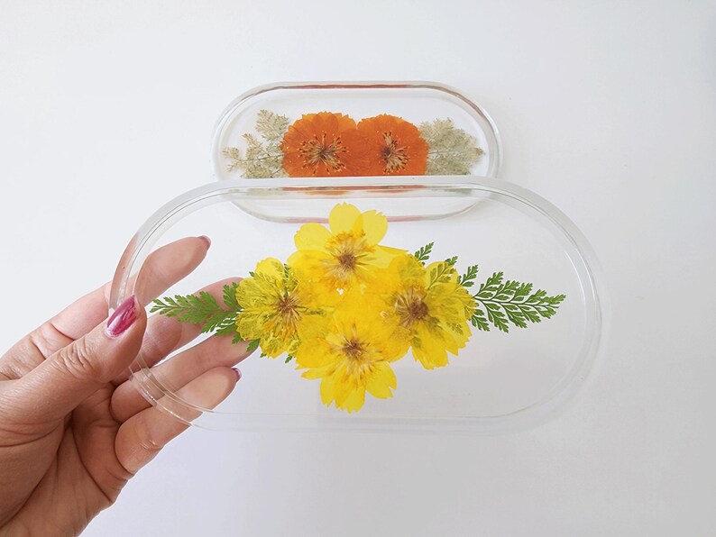May include: Two clear, oval-shaped resin trays. One tray features orange flowers, while the other holds yellow flowers and green fern-like leaves. The trays are displayed on a white background.