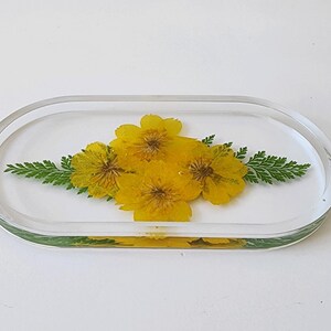 May include: Clear, oval-shaped resin tray with embedded yellow flowers and green fern-like leaves. The flowers are centered, and the leaves are on either side. The tray is on a white surface.