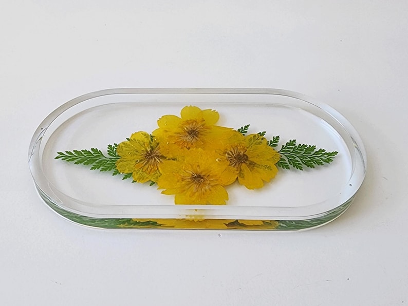May include: Clear, oval-shaped resin tray with embedded yellow flowers and green fern-like leaves. The flowers are centered, and the leaves are on either side. The tray is on a white surface.