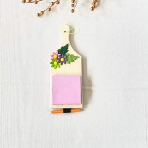 May include: A wooden memo board with a pink sticky note and a pencil. The board is decorated with a floral design and a green leaf.