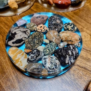 K&ouml;nnte beinhalten: Nahaufnahme eines blauen Harz-Untersetzers mit verschiedenen kleinen, bunten und gemusterten Steinen, die in das Harz eingebettet sind. Der Untersetzer befindet sich auf einer Holzoberfl&auml;che.
