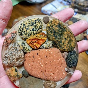 K&ouml;nnte beinhalten: Ein transparenter Harz-Untersetzer mit verschiedenen bunten und gemusterten Steinen, die darin eingebettet sind. Die Steine sind in verschiedenen Braunt&ouml;nen, Gr&uuml;n, Gelb und Rot.