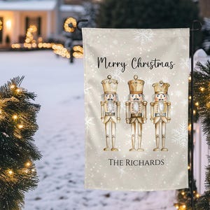 Puede incluir: Una bandera de jardín navideña beige con tres figuras de cascanueces con coronas y detalles dorados. La bandera dice "Merry Christmas" y "The Richards" en escritura negra, con detalles de copos de nieve. La bandera se muestra al aire libre en un entorno nevado.