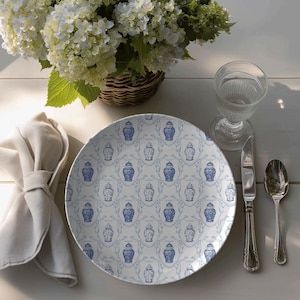 May include: A white round table set for dining outdoors. A plate with a blue and white pattern, a glass, silverware, and a napkin are arranged around a basket of white flowers.
