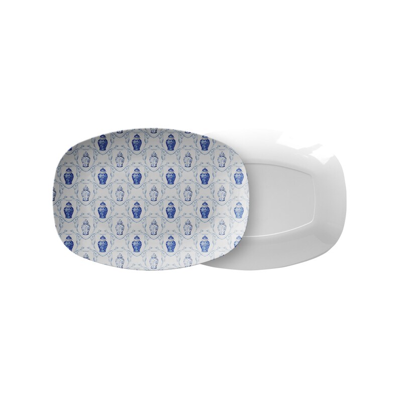May include: Two white rectangular serving platters. One platter features a blue and white pattern of decorative vases and floral elements. The other platter is plain white. The platters are designed for serving food.