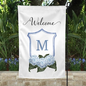 May include: A white welcome garden flag with a blue and white shield featuring the letter M. The shield is surrounded by blue hydrangeas with green leaves.