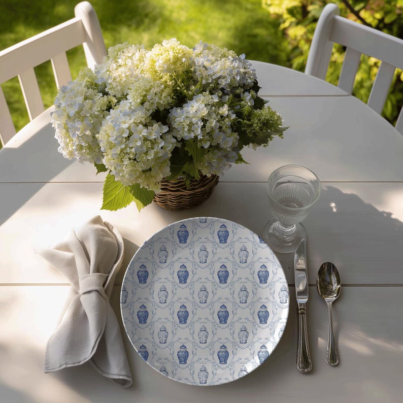 May include: A white round table set for dining outdoors. A plate with a blue and white pattern, a glass, silverware, and a napkin are arranged around a basket of white flowers.