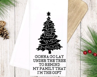 May include: A white kitchen towel with a black silhouette of a Christmas tree and the text "Gonna go lay under the tree to remind my family that I'm the gift."