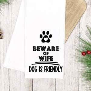 May include: White kitchen towel with black text that reads "Beware of Wife Dog is Friendly" with a paw print with a heart in the center.