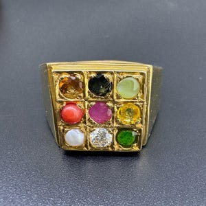 May include: A gold-toned ring featuring a square face with nine colorful gemstones. The gemstones include shades of orange, black, green, red, pink, yellow, white, and a clear diamond. The ring is set against a dark background.