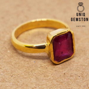 Op de afbeelding: Een gouden ring met een grote, vierkante, rode edelsteen. De ring is gezet in een eenvoudige, gouden band.