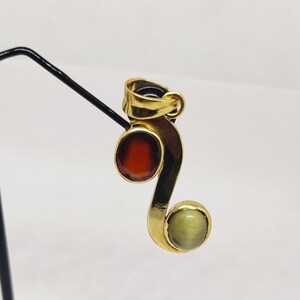 May include: A gold pendant with a red and green gemstone. The pendant is shaped like a sideways 'S' with a round red gemstone at the top and a round green gemstone at the bottom.