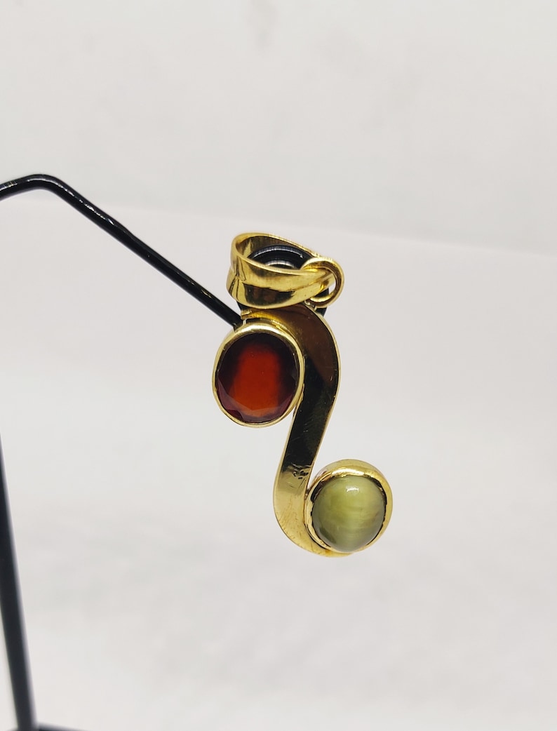 May include: A gold pendant with a red and green gemstone. The pendant is shaped like a sideways 'S' with a round red gemstone at the top and a round green gemstone at the bottom.