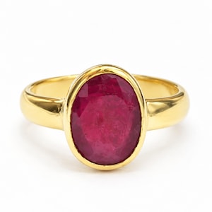 May include: A gold ring featuring a large, oval-cut, deep red gemstone set in a smooth, polished bezel setting. The band is solid gold with a rounded, high-shine finish, showcasing a classic design that highlights the vibrant color of the stone.
