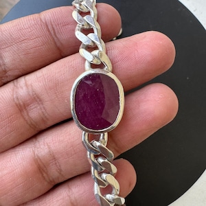Puede incluir: Un brazalete de cadena de plata con una piedra preciosa grande, de forma ovalada, de color rojo oscuro en el centro.