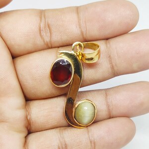 May include: Gold pendant with a red and a white gemstone. The pendant is shaped like a stylized letter S.