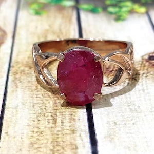 May include: A rose gold ring featuring an oval-cut ruby gemstone. The ruby is a deep red color and is held in place by four prongs. The band has an open, decorative design.
