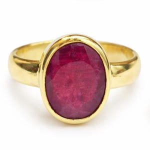 May include: A gold ring featuring a large, oval-cut ruby gemstone. The ruby is a deep, rich red color, set in a polished gold band. The ring's design is simple yet elegant, showcasing the gemstone's beauty.