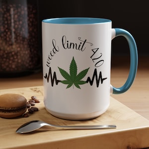 Puede incluir: Taza de cerámica blanca con borde y asa azules. La taza presenta una hoja de marihuana verde con un gráfico de latido del corazón y el texto "weed limit 420".