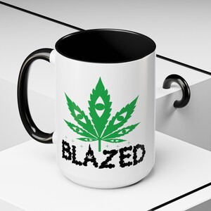 May include: White ceramic mug with a black handle and rim. The mug features a green marijuana leaf with eyes and the word "BLAZED" in black letters.