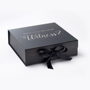 Puede incluir: Caja de regalo negra con lazo de cinta de raso negro a juego. La tapa presenta el texto "WILL YOU BE OUR Witness?" en plata. Ideal para un regalo especial o una petición de boda.