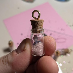May include: A small glass bottle with a cork stopper and a gold metal loop. The bottle is filled with pink glitter and a miniature figurine.