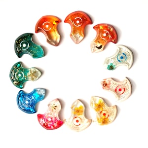 Puede incluir: Una colección de doce colgantes de resina en varios colores, dispuestos en círculo. Cada colgante presenta un diseño único con purpurina y elementos incrustados como flores y estrellas de mar. La paleta de colores incluye tonos de naranja, verde, azul y rosa.