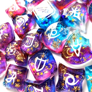 Op de afbeelding: Een set van 12-zijdige dobbelstenen met een galaxie thema. De dobbelstenen zijn gemaakt van helder hars met een wervelende mix van roze, blauwe en paarse kleuren. Elke dobbelsteen heeft een ander symbool erop, en de symbolen zijn wit omrand. De dobbelstenen zijn bedekt met gouden glitter.