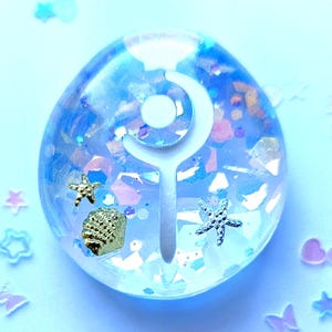 Op de afbeelding: Een heldere, ovale hars hanger met een wit, gestileerd ontwerp. De hanger is gevuld met blauwe en iriserende glitter, en bevat kleine gouden en zilveren zeesterren en een gouden zeeschelp. De achtergrond is lichtblauw.