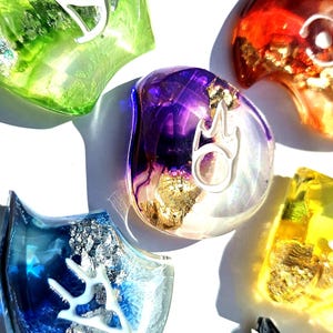 Op de afbeelding: Een assortiment kleurrijke, doorschijnende harsstukken met ingebedde metallic vlokken en witte symbolen. De stukken zijn in tinten groen, blauw, paars, rood en geel. De vormen variëren, sommige lijken op schilden en andere zijn ronder.