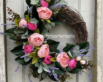 May include: A grapevine wreath adorned with pink peonies, fuchsia buds, and lavender sprigs. The wreath also features large green leaves and smaller gray-green leaves. The text "lavenderheartwreaths" is visible on the bottom of the wreath.
