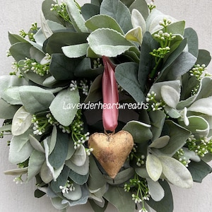May include: A green and white faux eucalyptus wreath with a gold heart ornament hanging from a pink ribbon. The wreath is decorated with small white flowers.