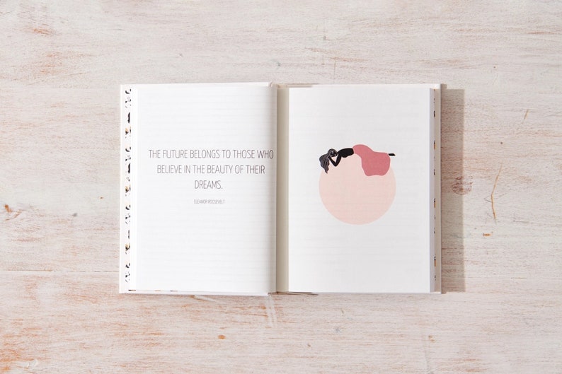 May include: An open journal with a white background and black lines. The left page has a quote that reads "The future belongs to those who believe in the beauty of their dreams." The right page has a simple illustration of a person lying on a pink circle.