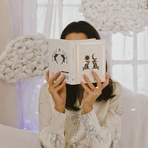 May include: A person is holding a white book open with a black and white illustration of two figures with a sun and moon above them. The book's title is "Dream Journal".